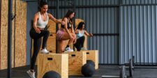Streamlining the WOD: Why CrossFit Studio Management Software Is Essential Three women performing box step-ups during a CrossFit workout inside a modern gym. Wooden plyo boxes, barbells, and medicine balls are visible on the floor, with metal panel walls in the background.