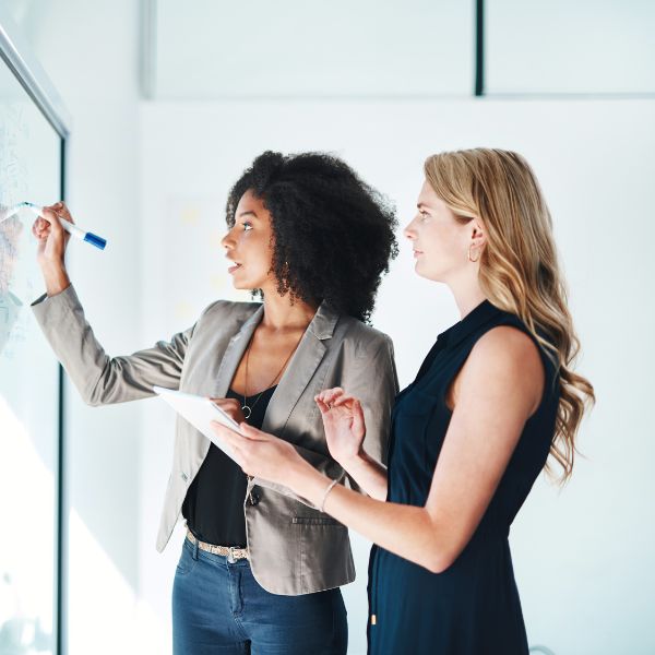 Two businesswomen collaborating at a whiteboard, one writing with a marker while the other holds a tablet and listens attentively during a strategy discussion.