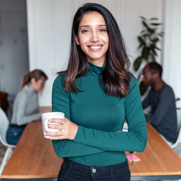 Smiling woman in a green turtleneck holding a coffee mug and standing confidently in a modern office, with two colleagues working in the background.