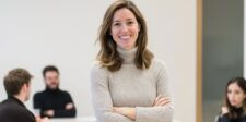 Smiling woman in a beige sweater stands with arms crossed in an office, with coworkers blurred in the background.