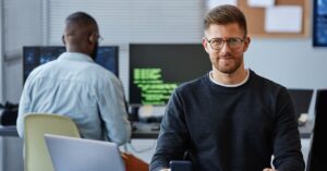 Two software developers working at their computers in a modern office, with one developer in glasses smiling at the camera, used as a hero image for a .NET 8 migration tutorial.
