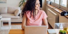 A smiling woman with curly hair sits at a wooden table using a laptop in a bright living room, looking out the window.