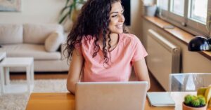 A smiling woman with curly hair sits at a wooden table using a laptop in a bright living room, looking out the window.