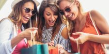 76 Psychological Factors in Marketing That Boost Sales Three young women wearing sunglasses smiling and leaning over shopping bags, looking excited about their purchases.