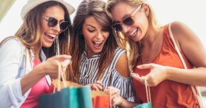 Three young women wearing sunglasses smiling and leaning over shopping bags, looking excited about their purchases.