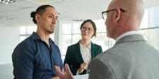Three business professionals having a discussion in a bright modern office, two men and one woman listening and speaking about a project or strategy.