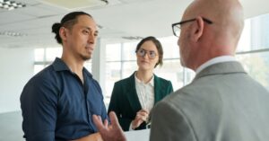 Three business professionals having a discussion in a bright modern office, two men and one woman listening and speaking about a project or strategy.