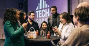 Group of attendees networking at a tech summit, talking around a table with drinks while “TECH SUMMIT” is displayed in the background.