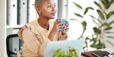 Woman sitting at her desk with a coffee mug, looking thoughtful while working on her laptop in a bright office.