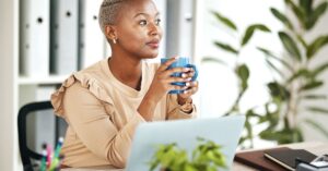 Woman sitting at her desk with a coffee mug, looking thoughtful while working on her laptop in a bright office.