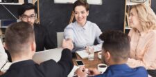 Team in a meeting room shaking hands across a table with laptops, phones, and coffee during a business discussion.