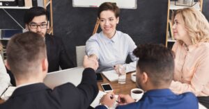 Team in a meeting room shaking hands across a table with laptops, phones, and coffee during a business discussion.
