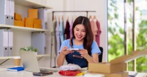 Smiling small business owner at her desk taking a product photo with her phone, surrounded by a laptop, parcels, and clothing in the background.