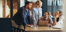 Four coworkers talk around a table with a laptop during an office meeting.
