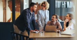 Four coworkers talk around a table with a laptop during an office meeting.