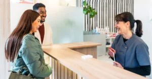 Receptionist smiling while helping two patients check in at a modern medical clinic front desk
