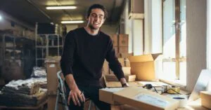 Small business owner in a warehouse workspace surrounded by packages and boxes, smiling while working at a packing table near a laptop.