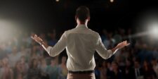 Speaker presenting from a podium with a laptop, facing a blurred audience in an auditorium.