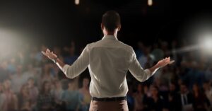Speaker presenting from a podium with a laptop, facing a blurred audience in an auditorium.