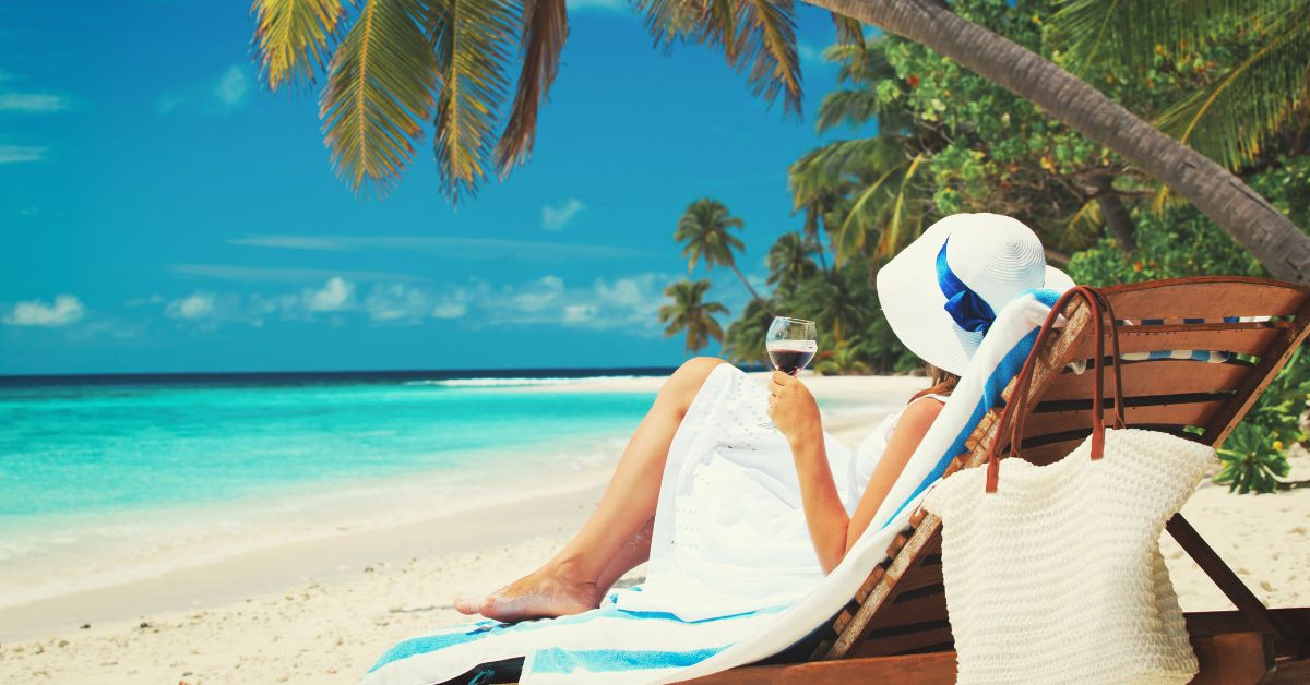 Person relaxing on a beach lounge chair under palm trees, holding a glass of wine by turquoise water.