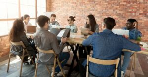 Team meeting around a table with laptops, discussing a project in a bright office.