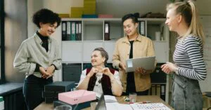 Four coworkers smiling and talking in an office meeting; one person holds a laptop while another sits at a table.