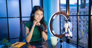 Woman recording a podcast at a desk with headphones, a microphone, a ring light, and a smartphone on a tripod.