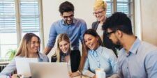 A group of coworkers gathered around a laptop in an office, smiling and looking at the screen during a team discussion.