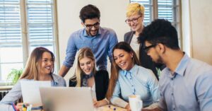 A group of coworkers gathered around a laptop in an office, smiling and looking at the screen during a team discussion.