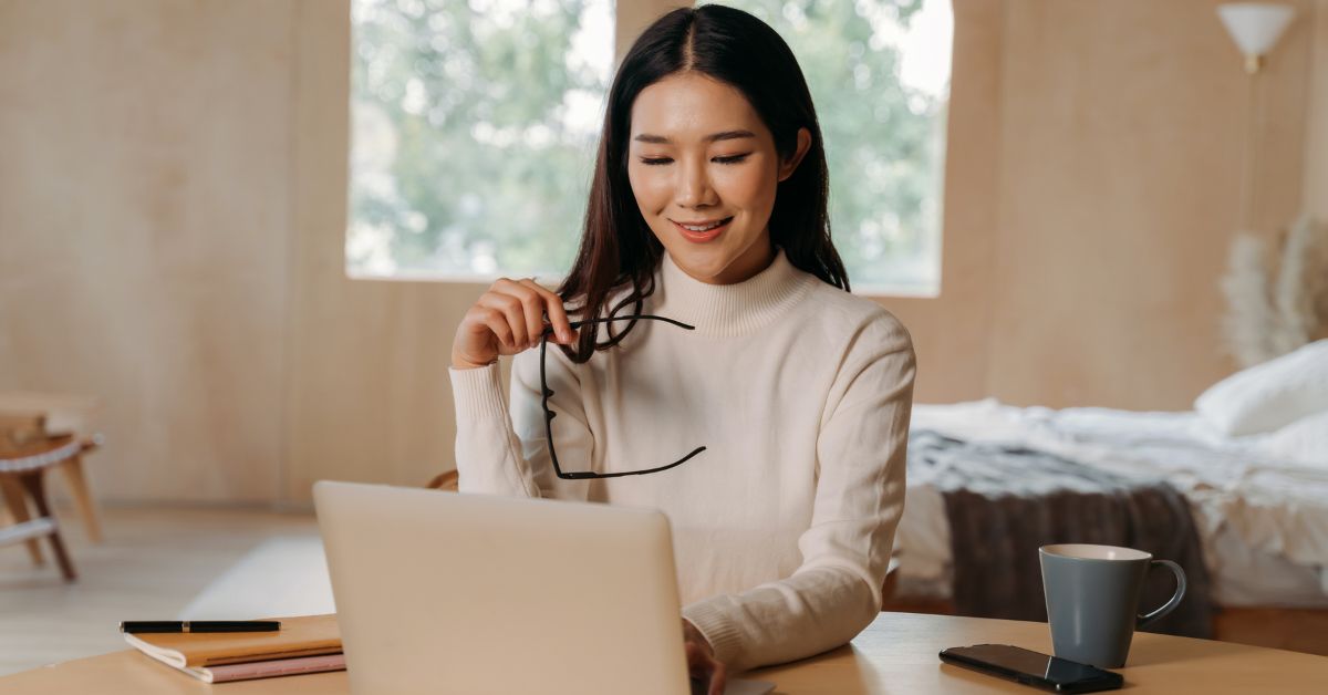 Woman working on a laptop while holding eyeglasses at a desk