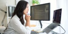 Person seated at a tidy workspace, focused on a large monitor displaying code, with a laptop on a stand and a mouse on the desk.