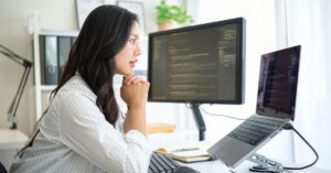 Person seated at a tidy workspace, focused on a large monitor displaying code, with a laptop on a stand and a mouse on the desk.