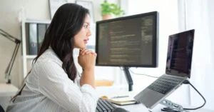 Person seated at a tidy workspace, focused on a large monitor displaying code, with a laptop on a stand and a mouse on the desk.