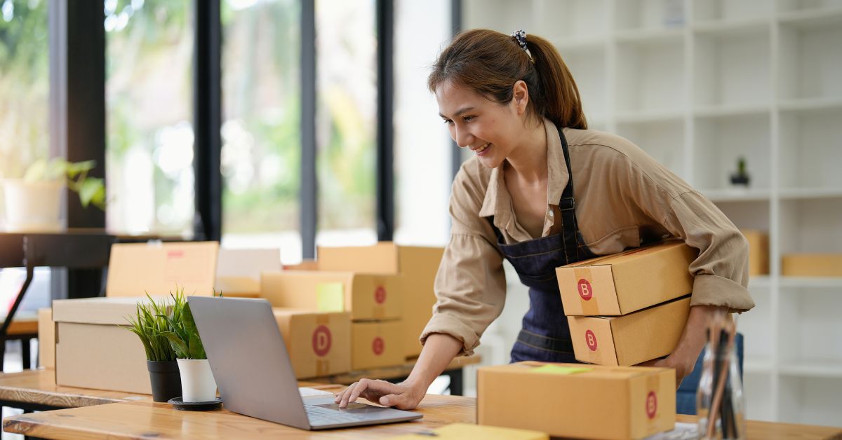Small business owner smiles while packing shipping boxes and working on a laptop in a bright workspace.