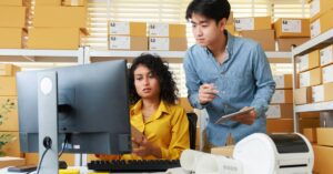 Two small business workers reviewing a computer screen in a warehouse office with shipping boxes behind them.