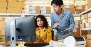Two small business workers reviewing a computer screen in a warehouse office with shipping boxes behind them.