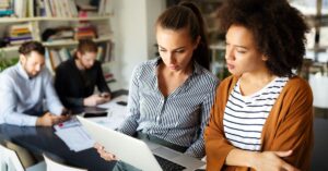 Two coworkers looking at a laptop together in an office while others work in the background