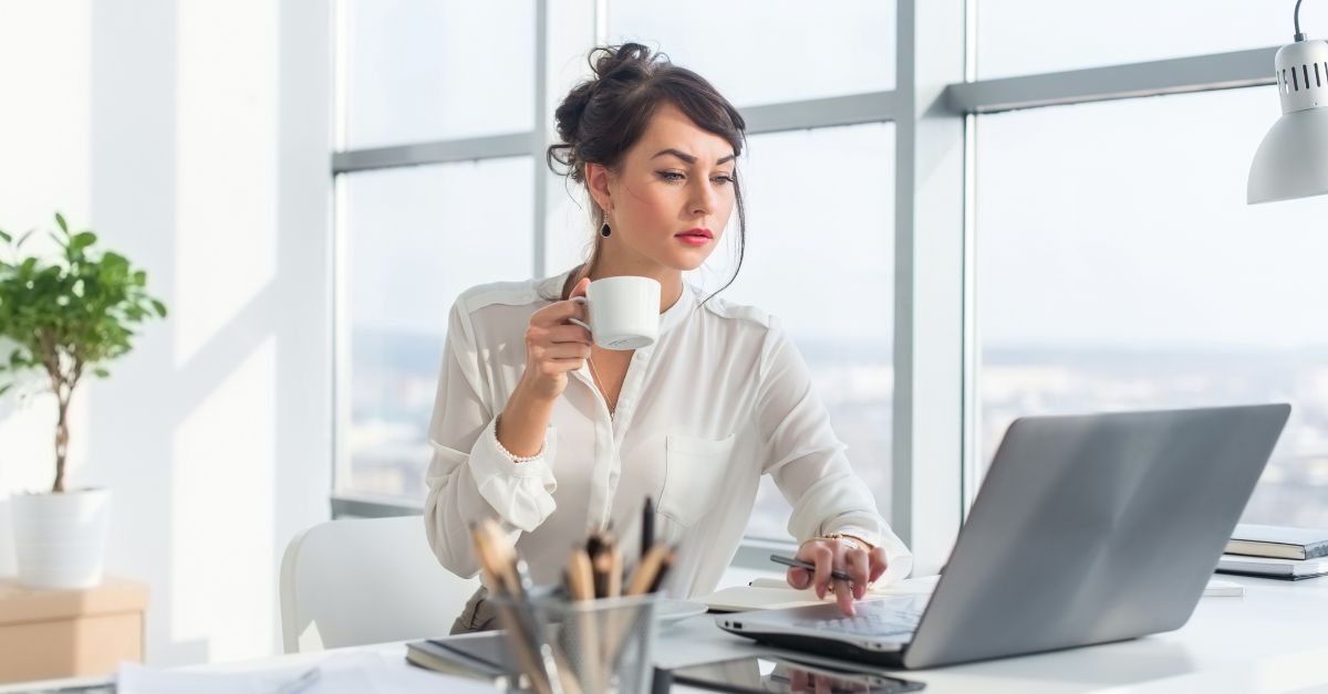 Woman in a bright office holding a coffee mug while looking at a laptop on her desk near large windows.