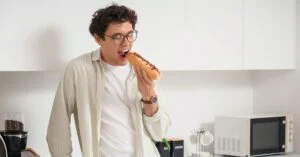 Man in a kitchen about to take a bite of a hot dog