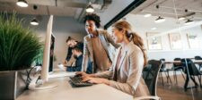 Two professionals review work at a desktop computer in a modern office while coworkers collaborate in the background.