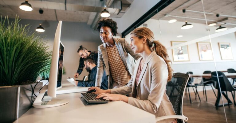 Two professionals review work at a desktop computer in a modern office while coworkers collaborate in the background.