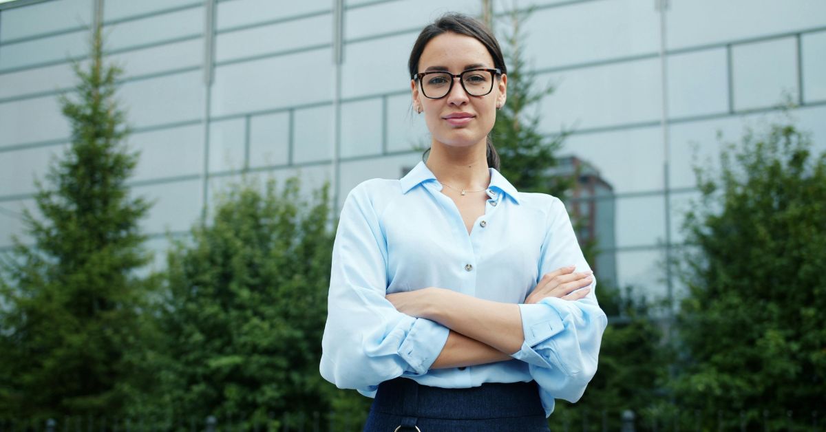 Professional woman in glasses and a light blue button-down shirt standing with arms crossed in front of a modern office building with green landscaping.