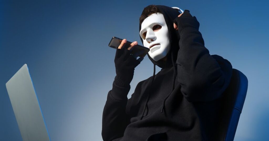 Masked person in a black hoodie holding a phone beside an open laptop, symbolizing AI-powered fraud or agentic fraud.