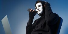 Masked person in a black hoodie holding a phone beside an open laptop, symbolizing AI-powered fraud or agentic fraud.