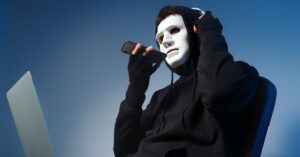 Masked person in a black hoodie holding a phone beside an open laptop, symbolizing AI-powered fraud or agentic fraud.