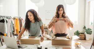 Two small business owners packing product boxes beside a laptop in a bright retail workspace, representing Canadian SMEs preparing orders while exploring business tools and AI adoption.