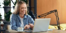 Woman speaking into a podcast microphone while working on a laptop at a desk with a coffee cup, notebook, and plants in the background.