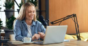 Woman speaking into a podcast microphone while working on a laptop at a desk with a coffee cup, notebook, and plants in the background.