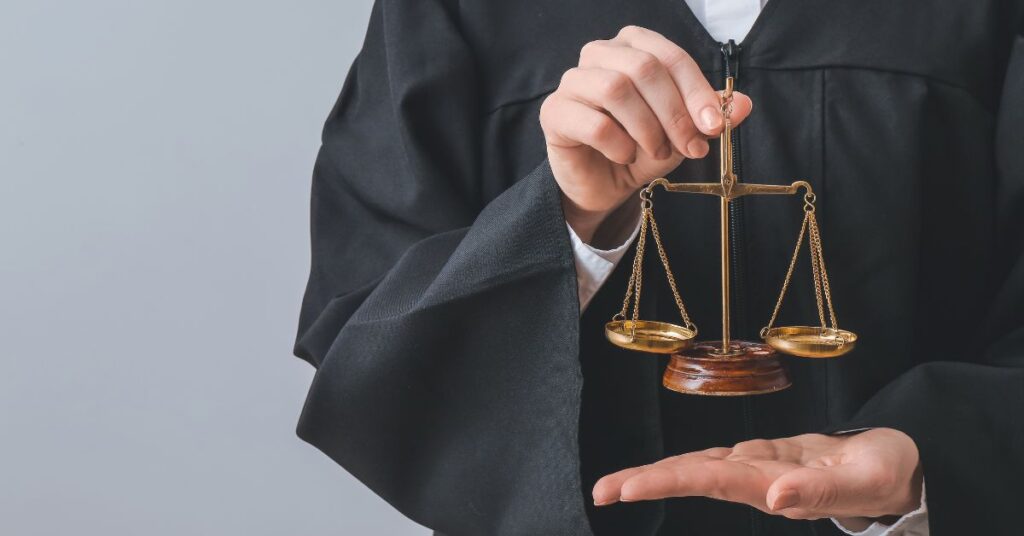 Person in a black judicial robe holding a small set of balanced gold scales of justice in an outstretched hand.