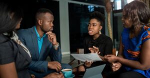 Four business professionals discussing a document during a meeting, with a laptop and coffee on the table.